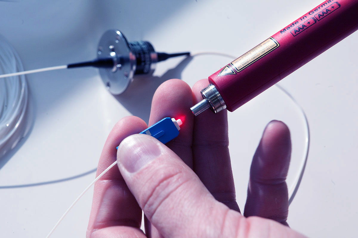 Hand hält einen Glasfaserstecker vor eine rote Lichtquelle.